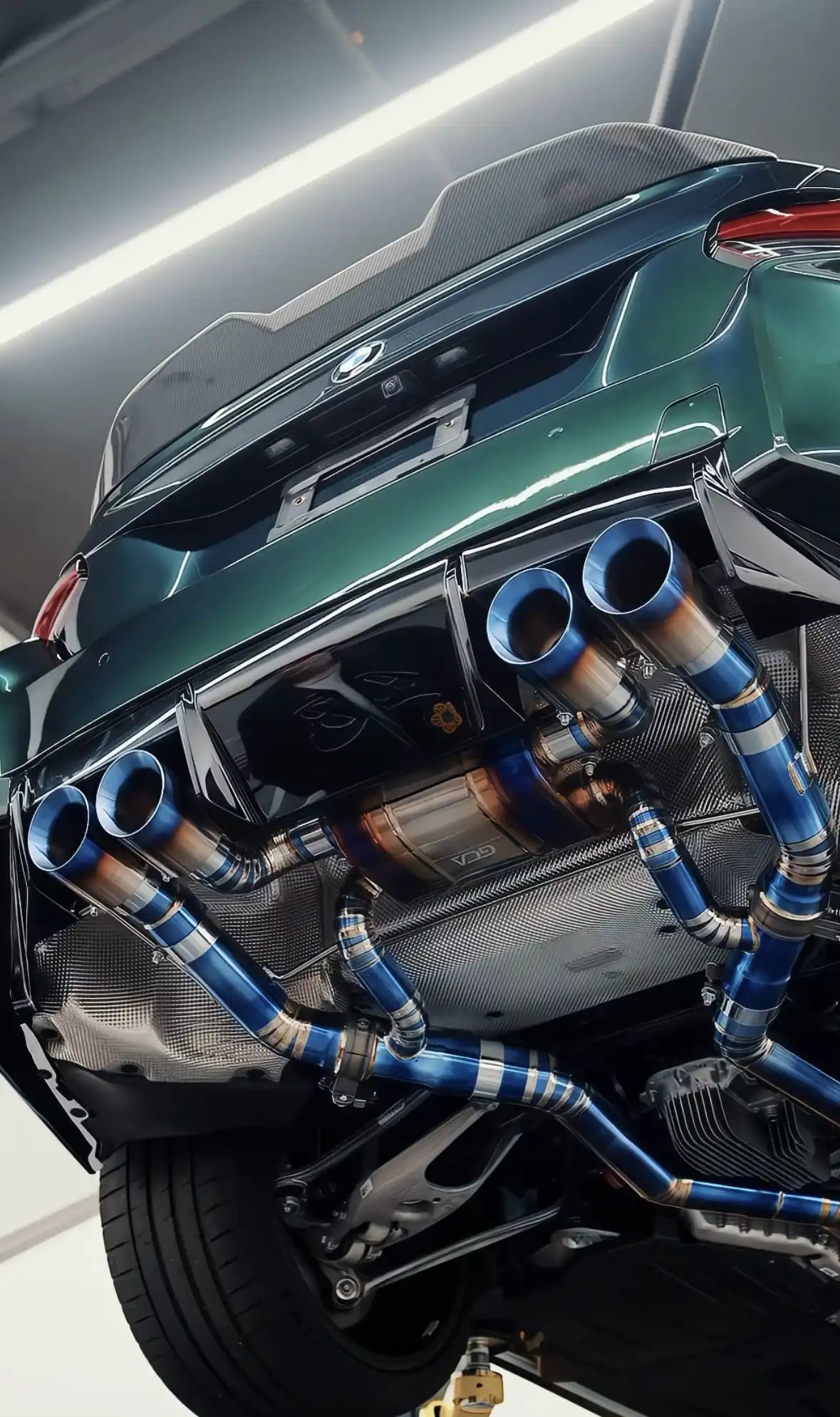 An aftermarket titanium exhaust system with blue-tinted tips and piping, installed on a dark green sports car.